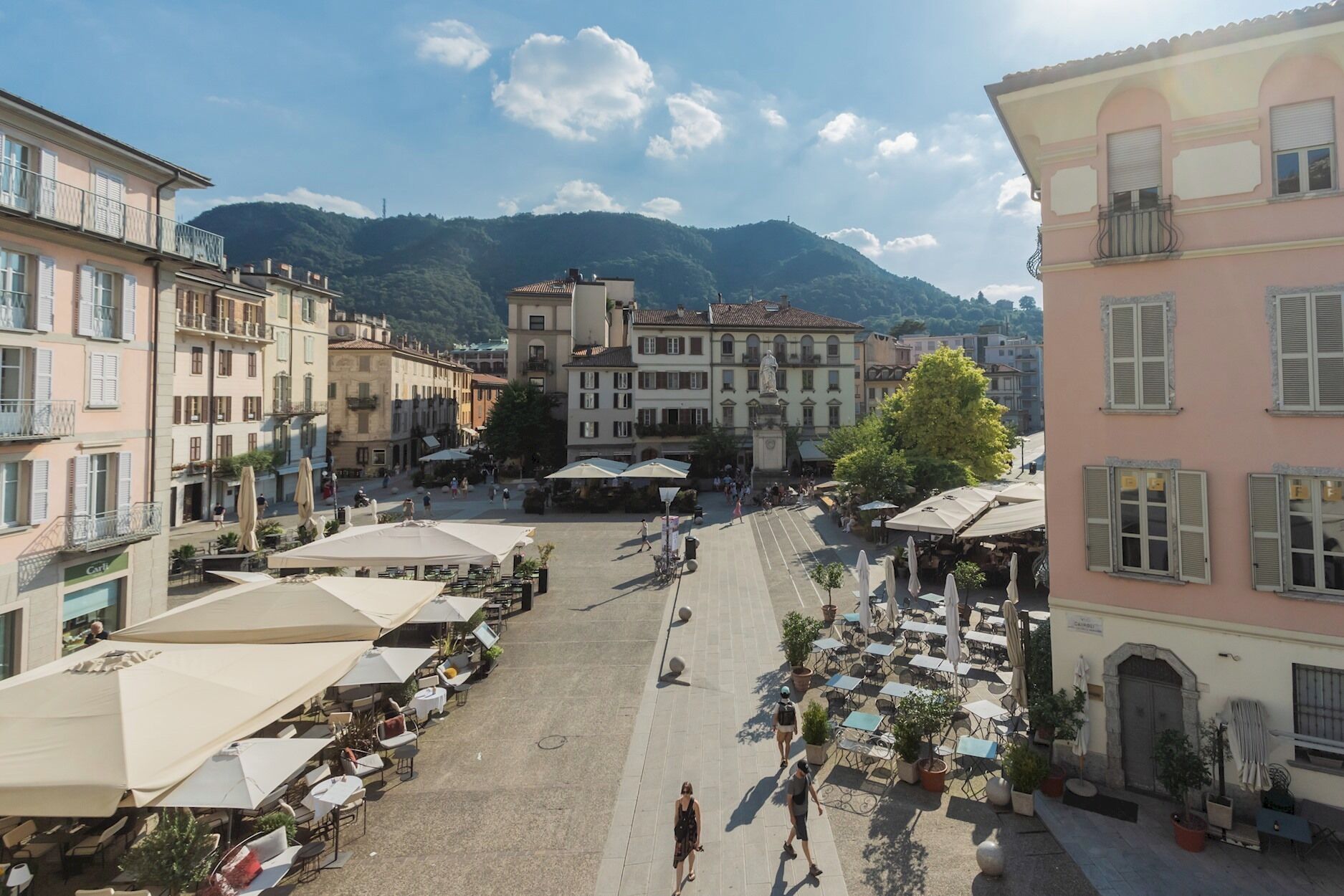 Una plaza con mesas al aire libre y una fuente, rodeada de edificios con balcones y una montaña al fondo.