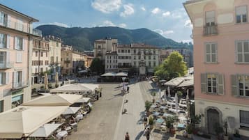 Una plaza con mesas al aire libre y una fuente, rodeada de edificios con balcones y una montaña al fondo.