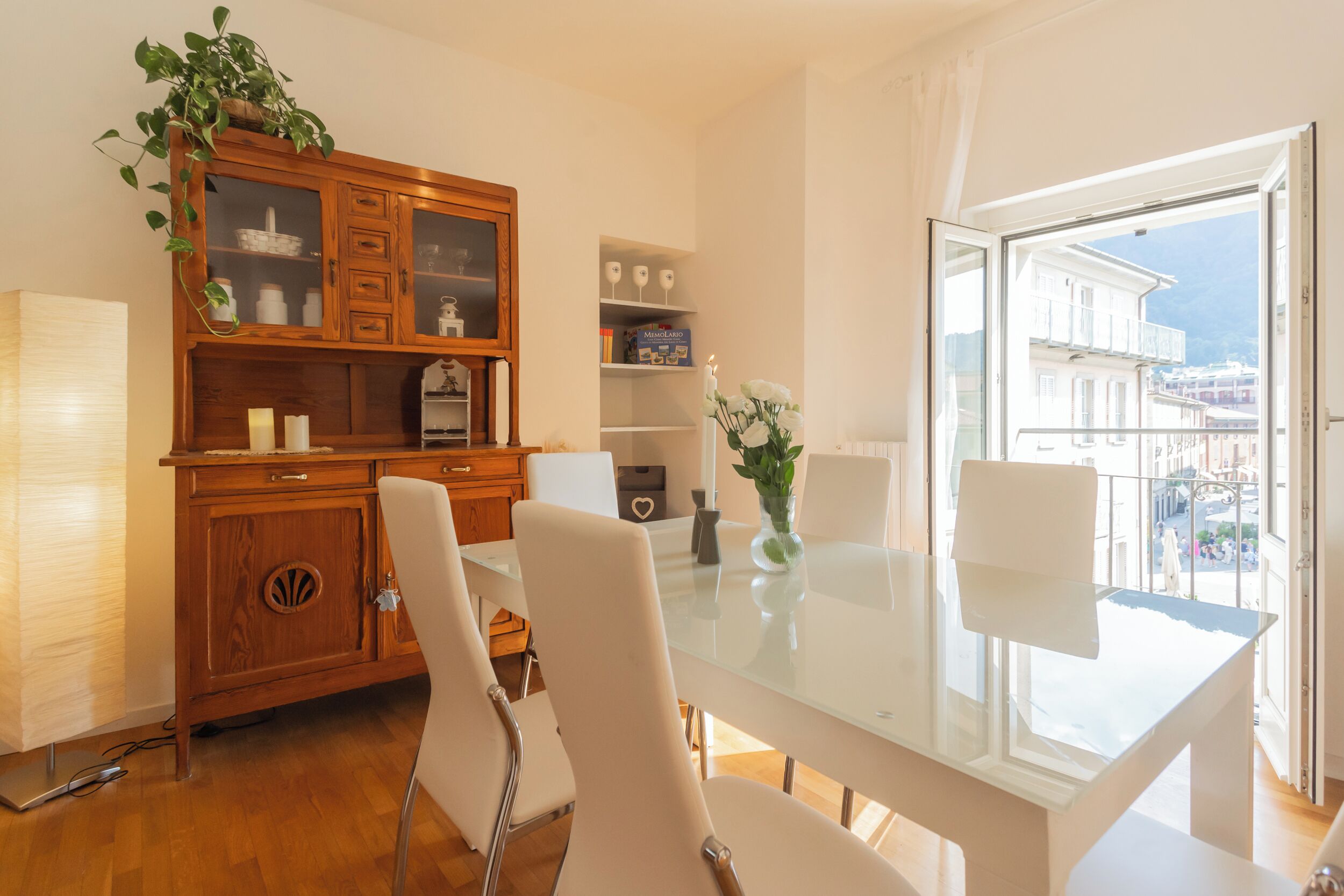 A modern dining area with a glass table, white chairs, a wooden cabinet with glass doors, and a view of buildings through a large window.