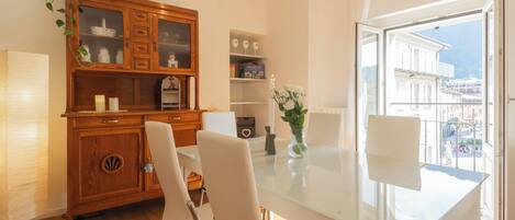 A modern dining area with a glass table, white chairs, a wooden cabinet with glass doors, and a view of buildings through a large window.