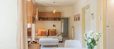 Una sala moderna con sofá, reposapiés y mesa de comedor. Se observa una zona de cocina con armarios de madera y una lámpara colgante.