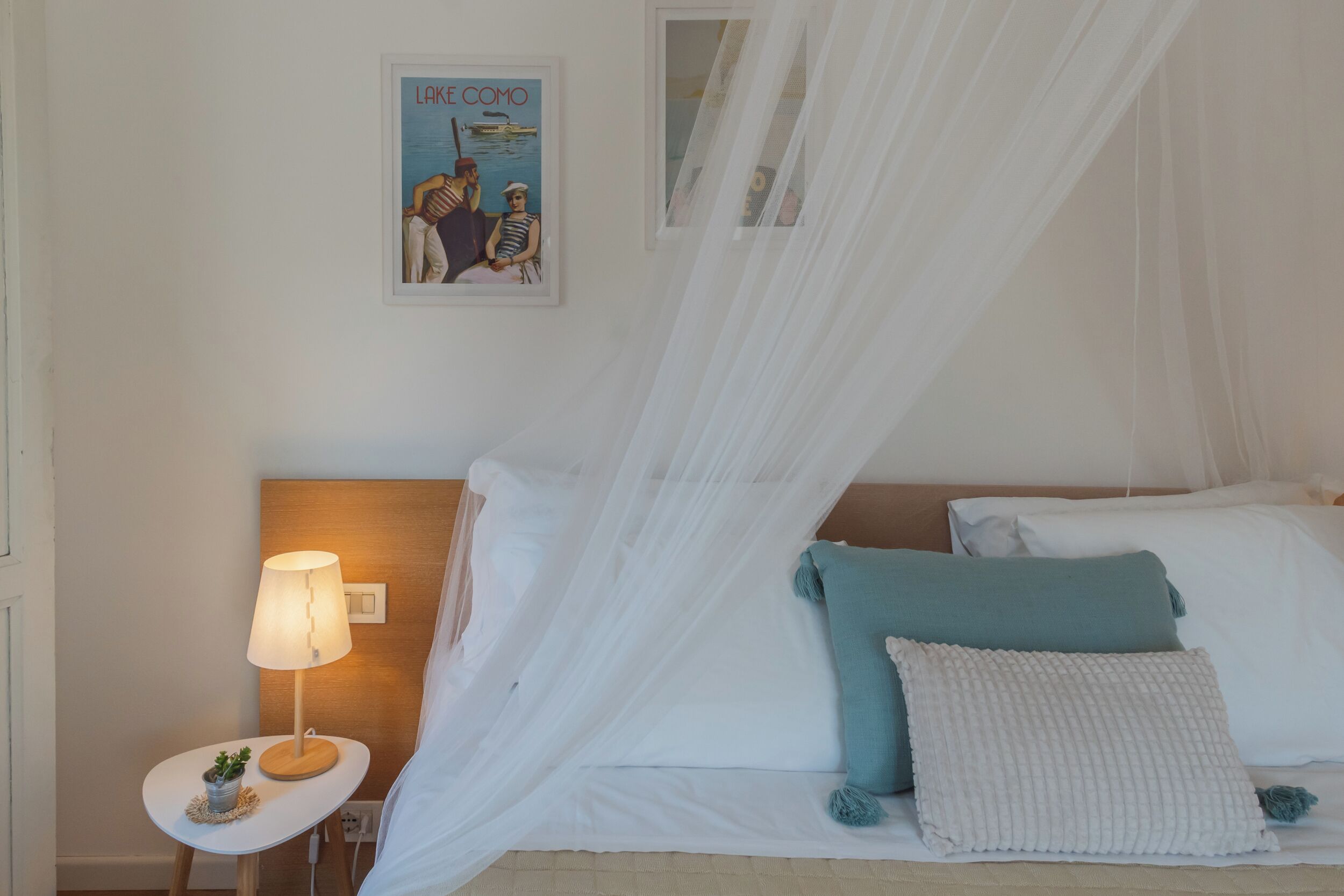 A neatly arranged bedroom with a bed, a bedside table with a lamp, a framed picture on the wall, and a sheer canopy over the bed.
