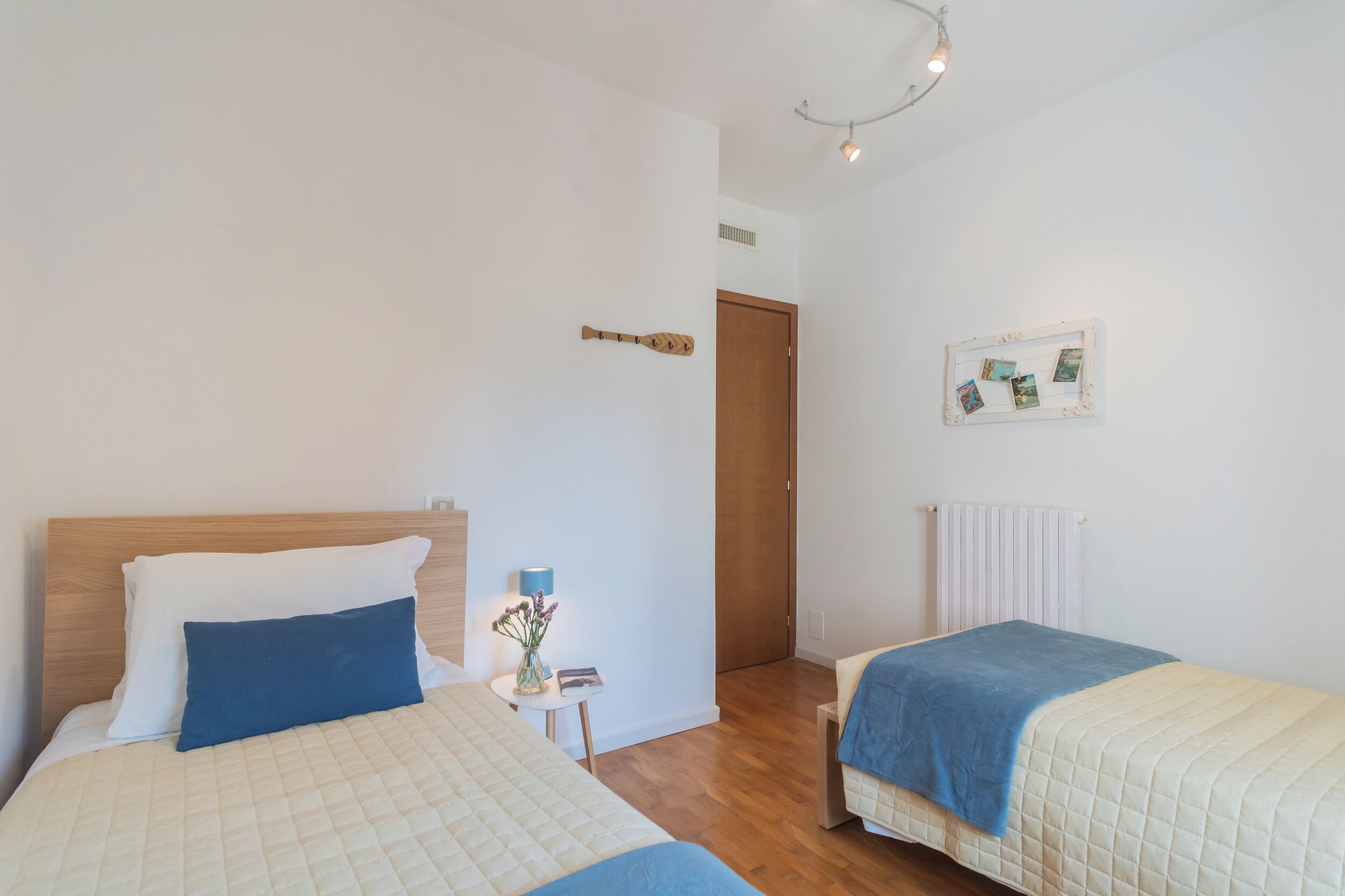 A bedroom with two beds, a wooden headboard, a small bedside table with a lamp, and a framed photo on the wall.