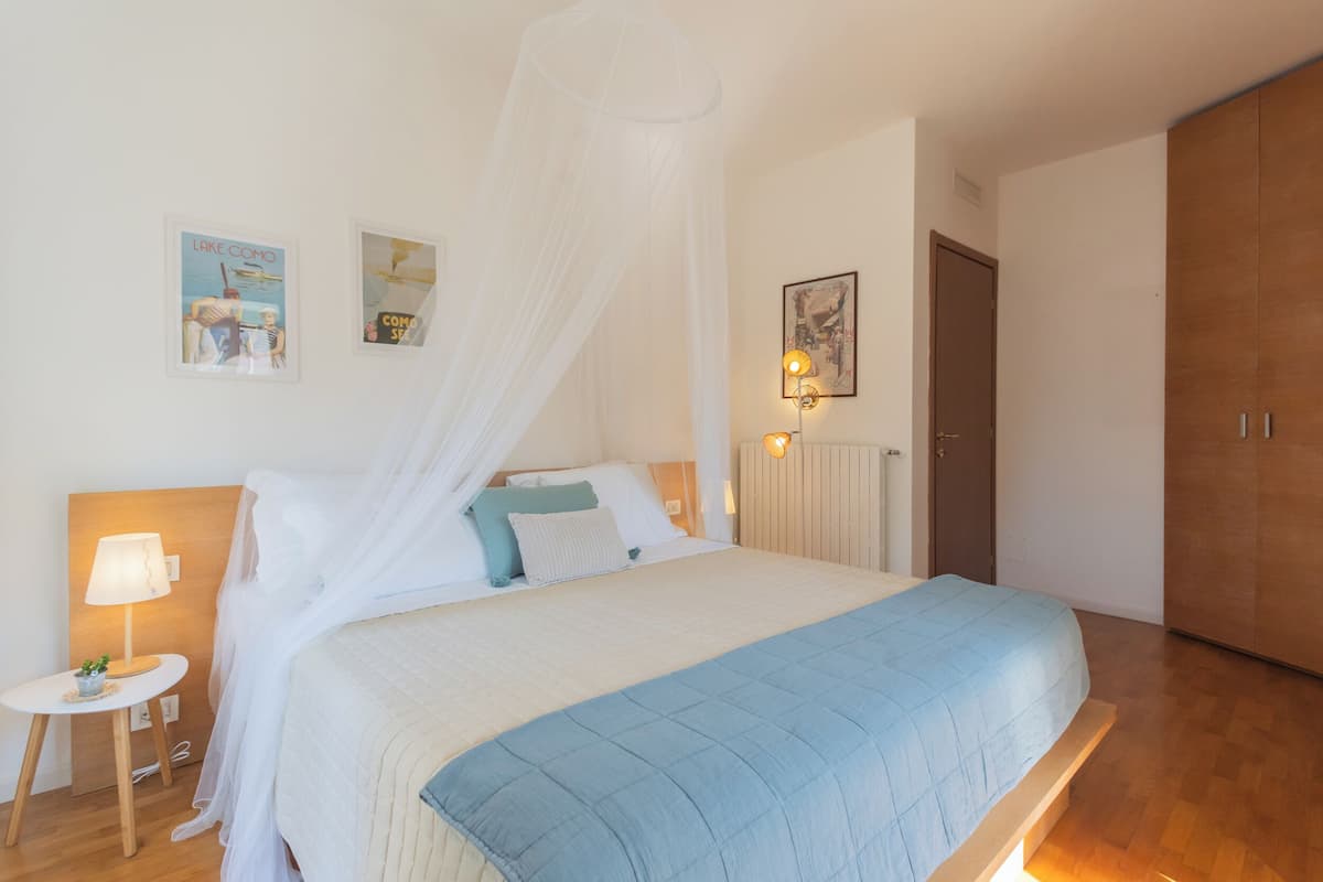 Un dormitorio con cama, mesita de noche con lámpara, luz en la pared y una puerta.