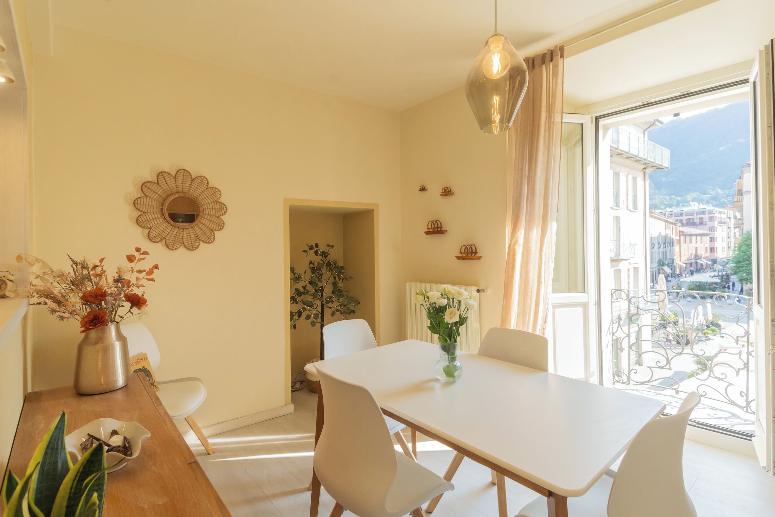 A dining area with a table and chairs, a vase with flowers, and a view of buildings through a balcony.