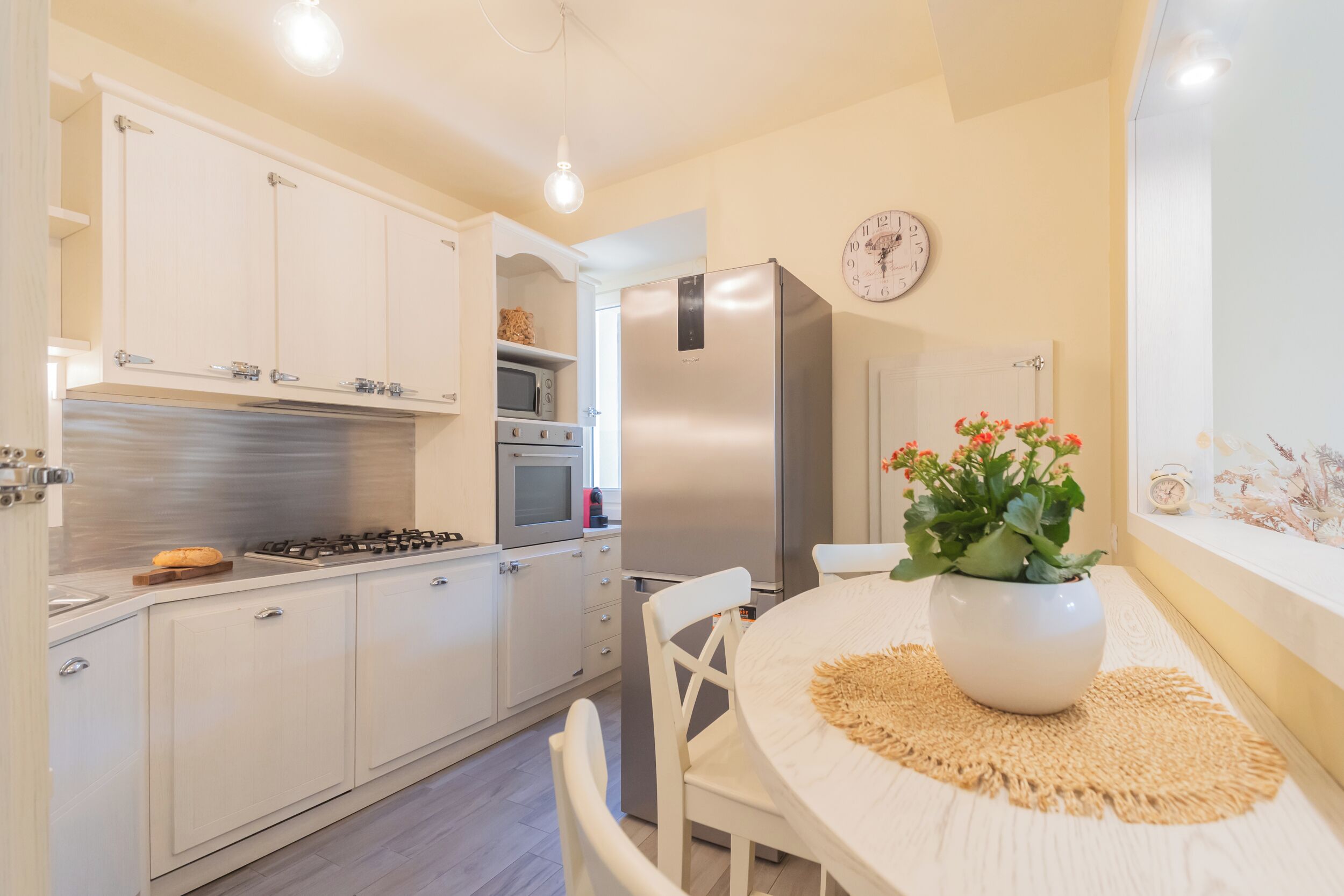 A modern kitchen with white cabinets, a stainless steel refrigerator, a microwave, and a gas stove. There is a round table with a white tablecloth and a potted plant.