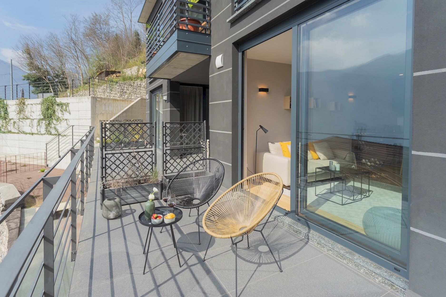 A modern balcony with a glass door, two wicker chairs, a small table with food and drink, and a view of the mountains.