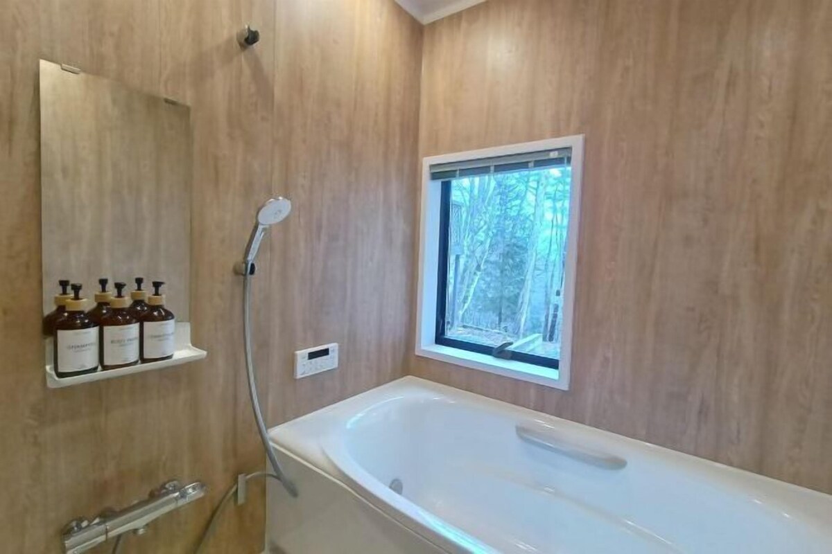 A modern bathroom with a freestanding bathtub, wooden panel walls, a window with a view of trees, and a shelf with bottles.
