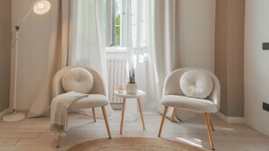 Ein modernes Zimmer mit zwei weißen Polsterstühlen, einem kleinen runden Tisch mit Pflanze, einer Stehlampe und durchsichtigen Vorhängen.