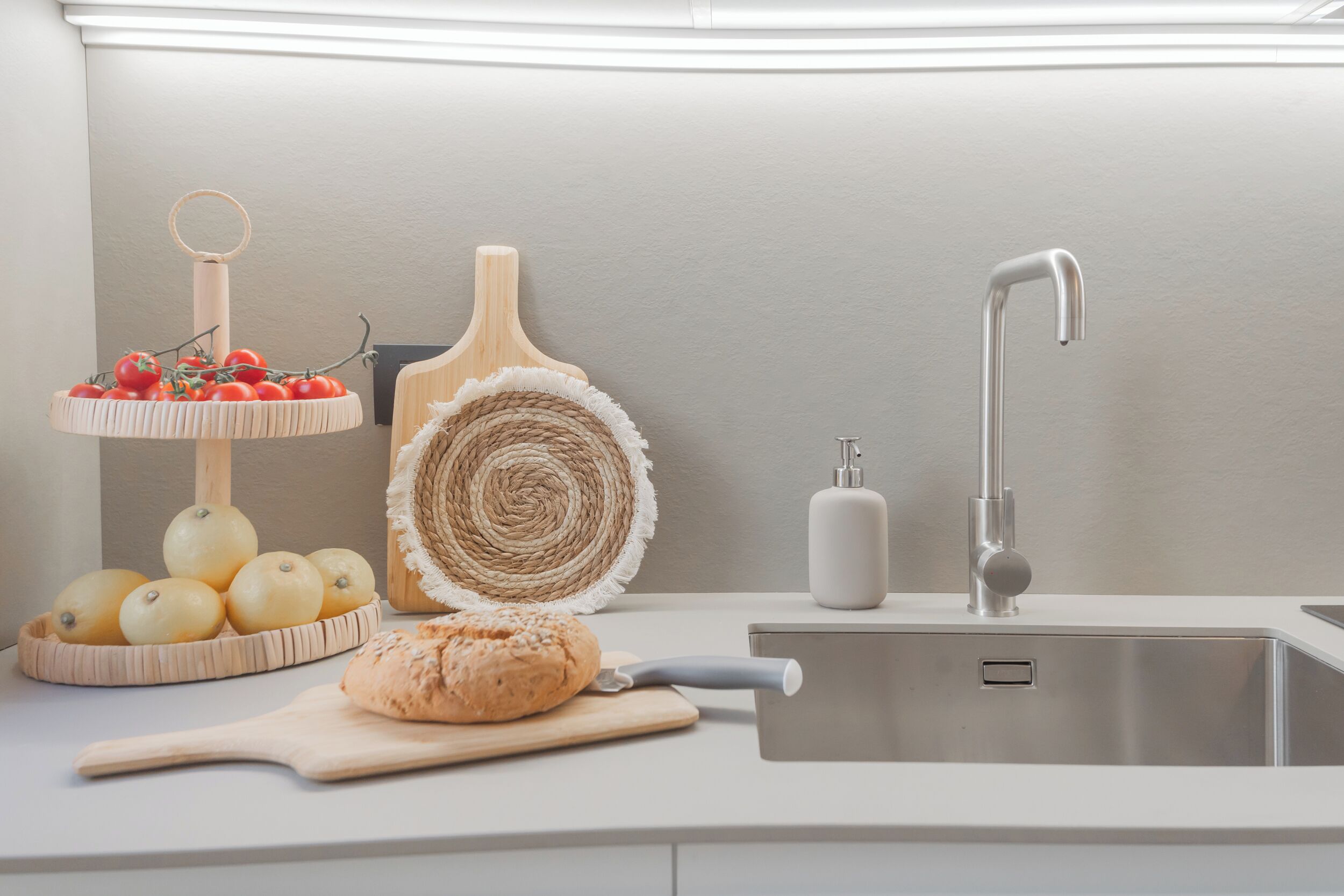 A kitchen counter with a sink, a bread loaf, a cutting board, a fruit basket, and a decorative wooden item.