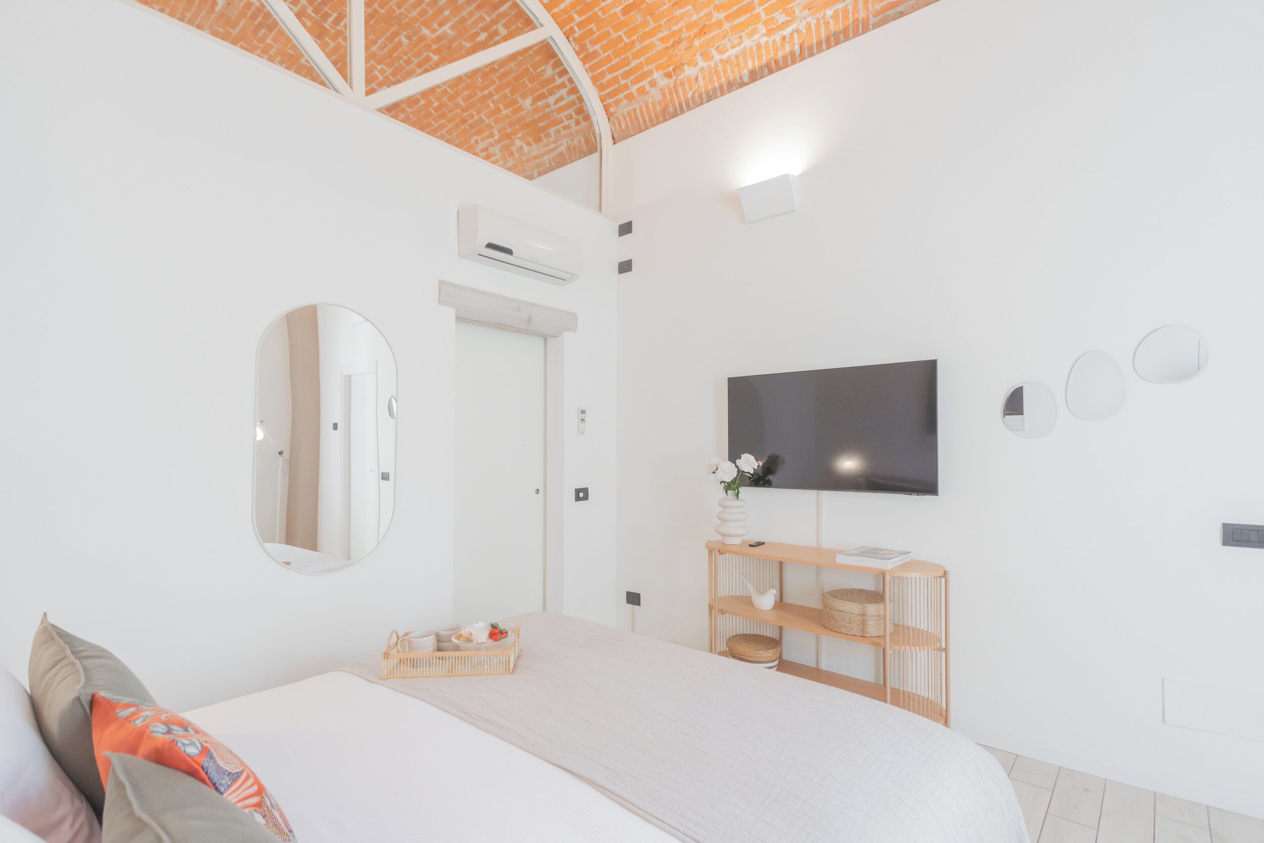 Una habitación de hotel moderna con una cama grande, un televisor de pantalla plana, un espejo, un estante de madera con cestas y un techo de ladrillo.