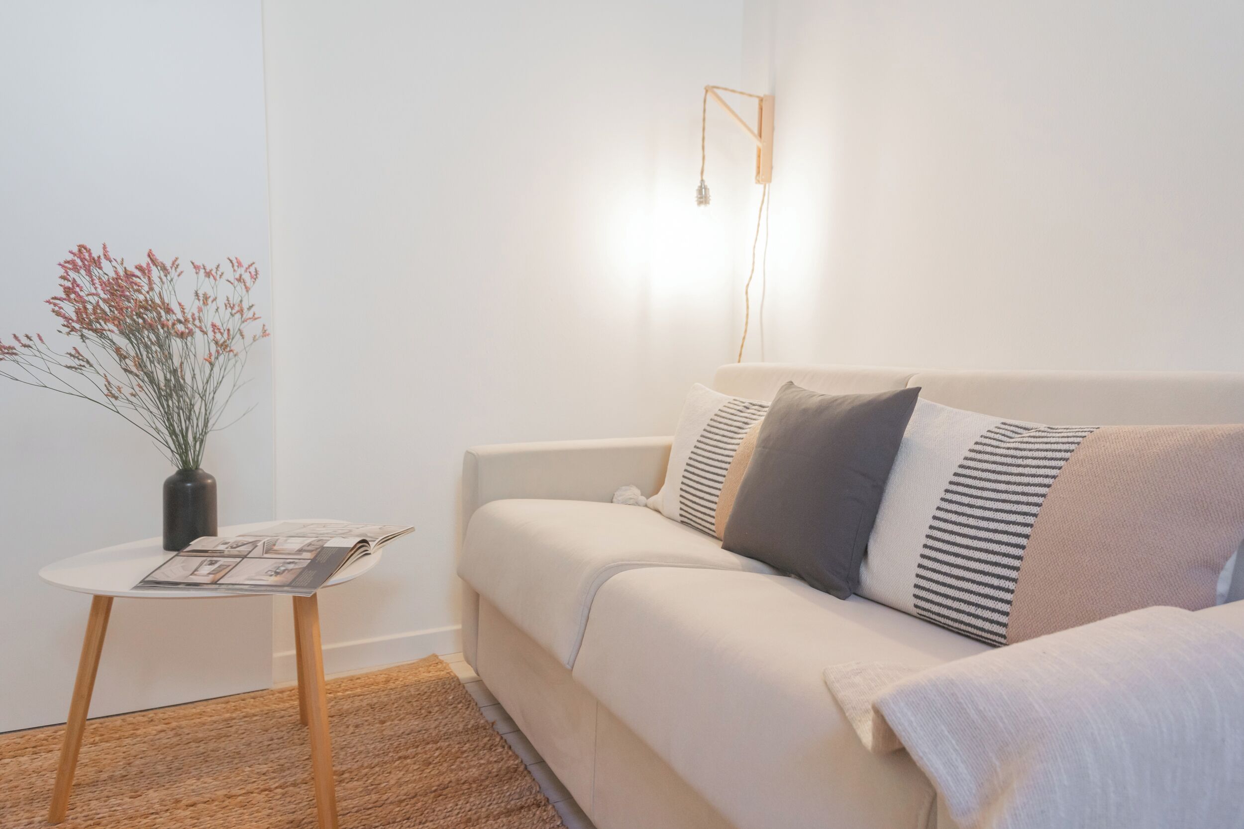 Una sala de estar minimalista con un sofá blanco, una mesita redonda con una revista, un jarrón con flores secas y una lámpara de pared.