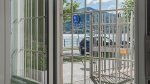 Blick durch ein Metallgitter, mit einem blauen Fußgängerzeichen, einer gepflasterten Fläche und einem Gewässer im Hintergrund.