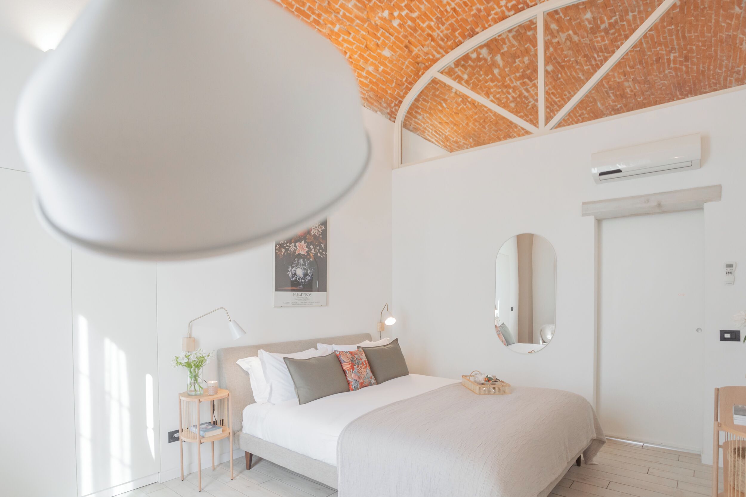 Un dormitorio moderno con techo de ladrillo, una cama con cabecera blanca, una mesita de noche con lámpara y un aire acondicionado instalado en la pared.