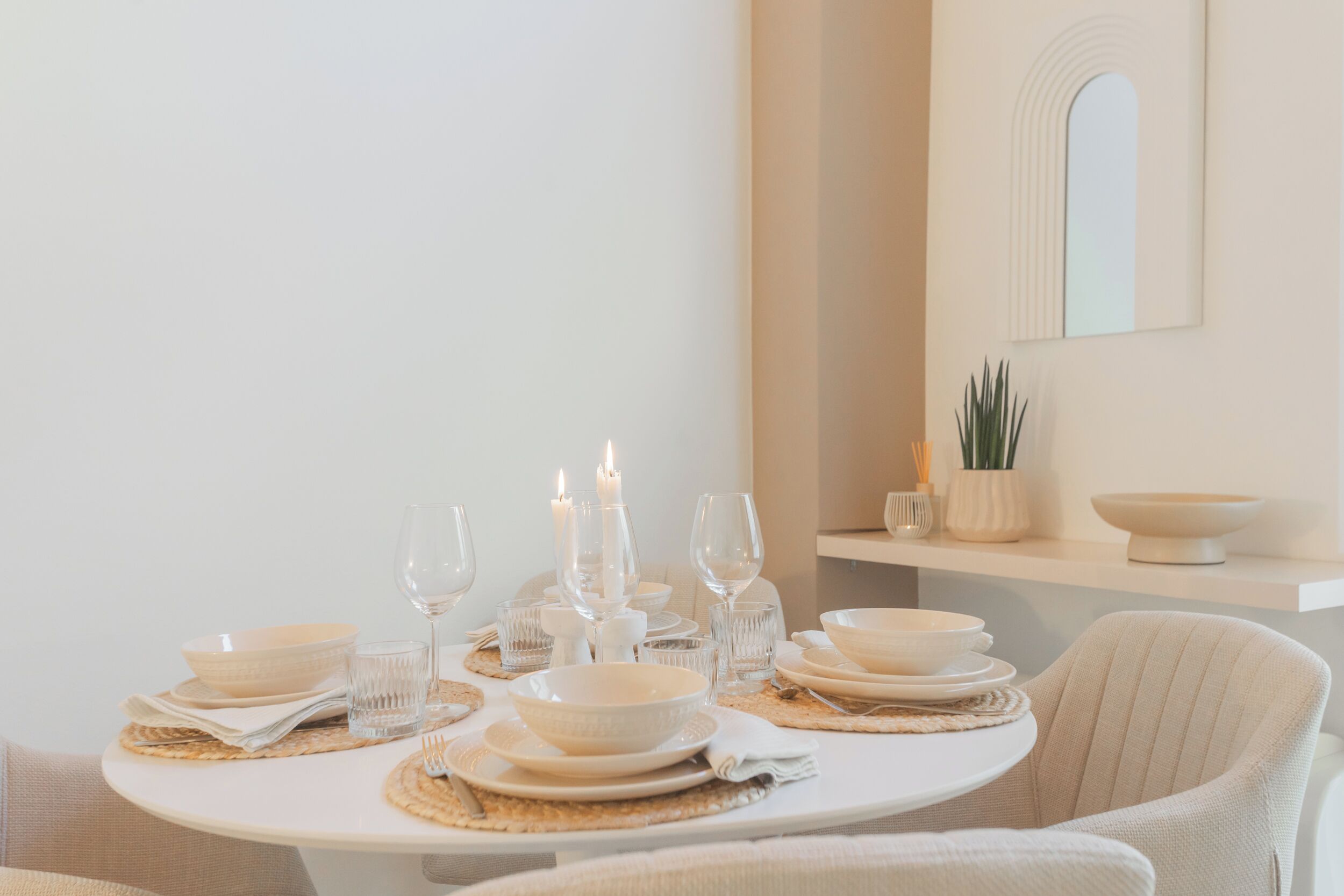 Una mesa de comedor dispuesta con platos, cuencos y vasos blancos, velas y un espejo en la pared.