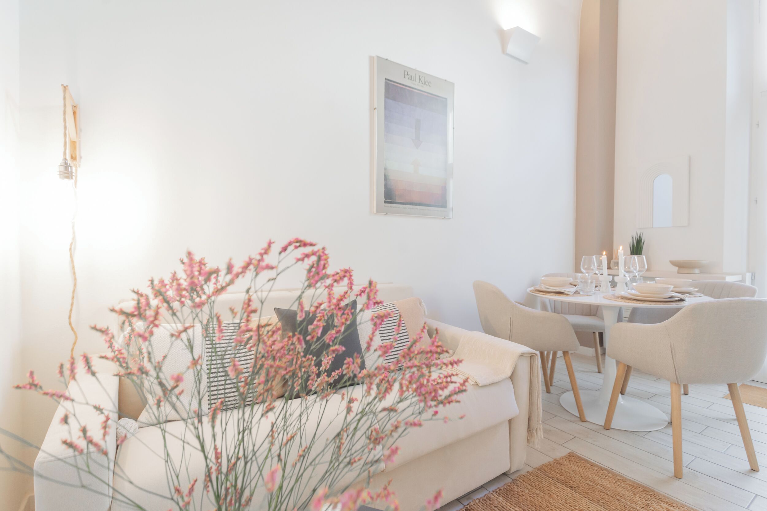 Een moderne woonkamer met een bank, eettafel en stoelen. Er hangt een kunstwerk aan de muur en op de bank staat een vaas met bloemen.