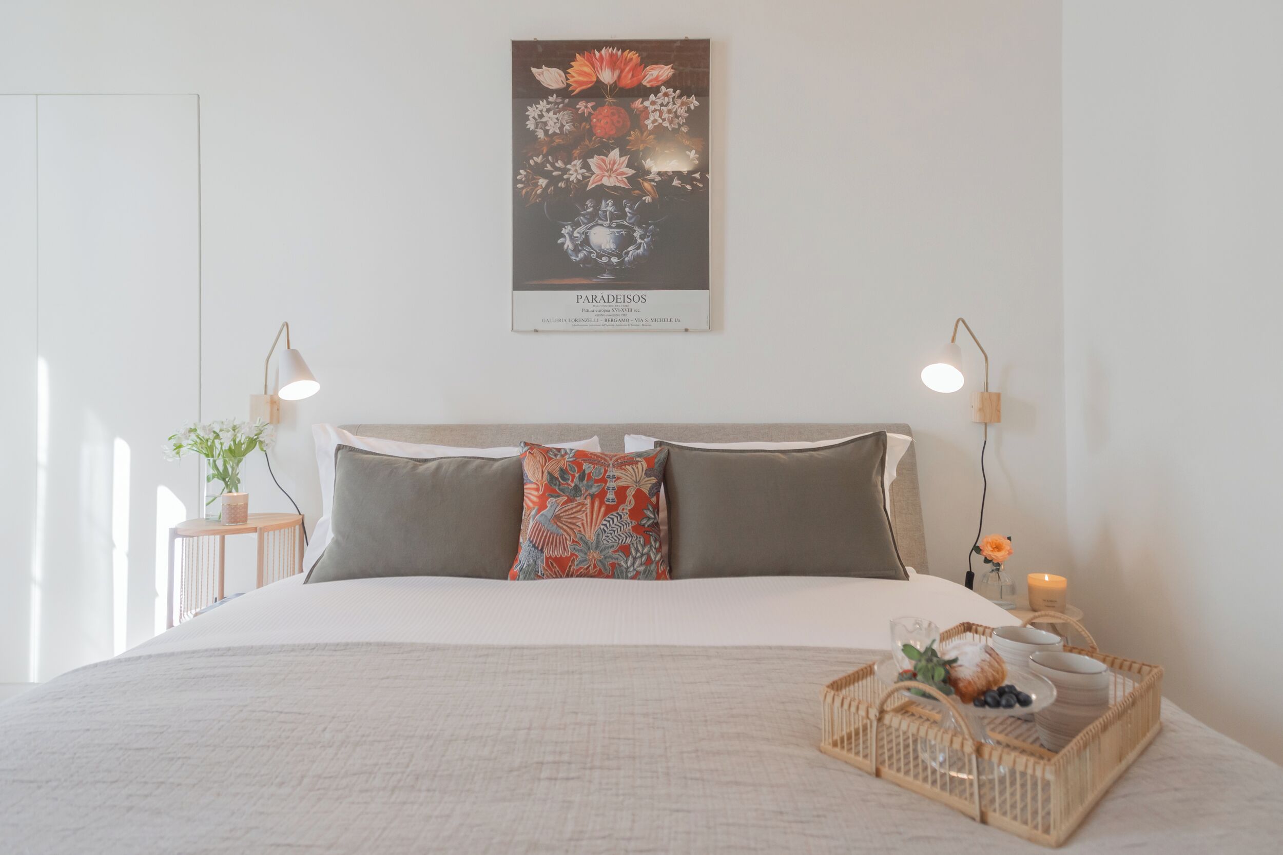A neatly arranged bedroom with a large bed, a decorative poster, bedside lamps, and a tray with fruits and a candle.