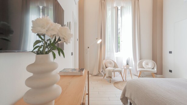 Ein modernes Schlafzimmer mit einem großen Bett, einer weißen Vase mit Blumen, zwei weißen Stühlen und einem kleinen Tisch mit einer Lampe.