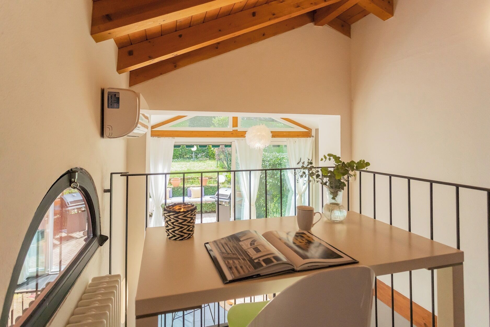 Ein Balkon mit Holzgeländer, einem Tisch mit einer Zeitschrift, einer Pflanze im Blumenvasen und Blick auf grüne Vegetation.
