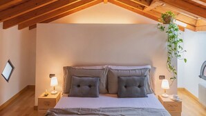 A bedroom with a sloped wooden ceiling, a bed with grey bedding, two bedside tables with lamps, and a hanging plant. - Vico Garden-203 by Rent all Como (Como)