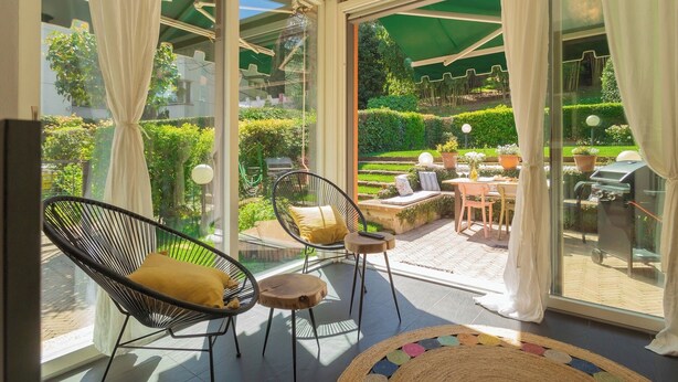 A sunlit room with large glass doors leading to an outdoor patio. Inside, there are two wicker chairs with cushions, a small round table, and a colorful woven rug. - Vico Garden-203 by Rent all Como (Como)