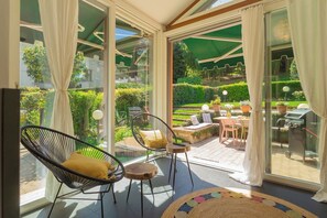 A sunlit room with large glass doors leading to an outdoor patio. Inside, there are two wicker chairs with cushions, a small round table, and a colorful woven rug.