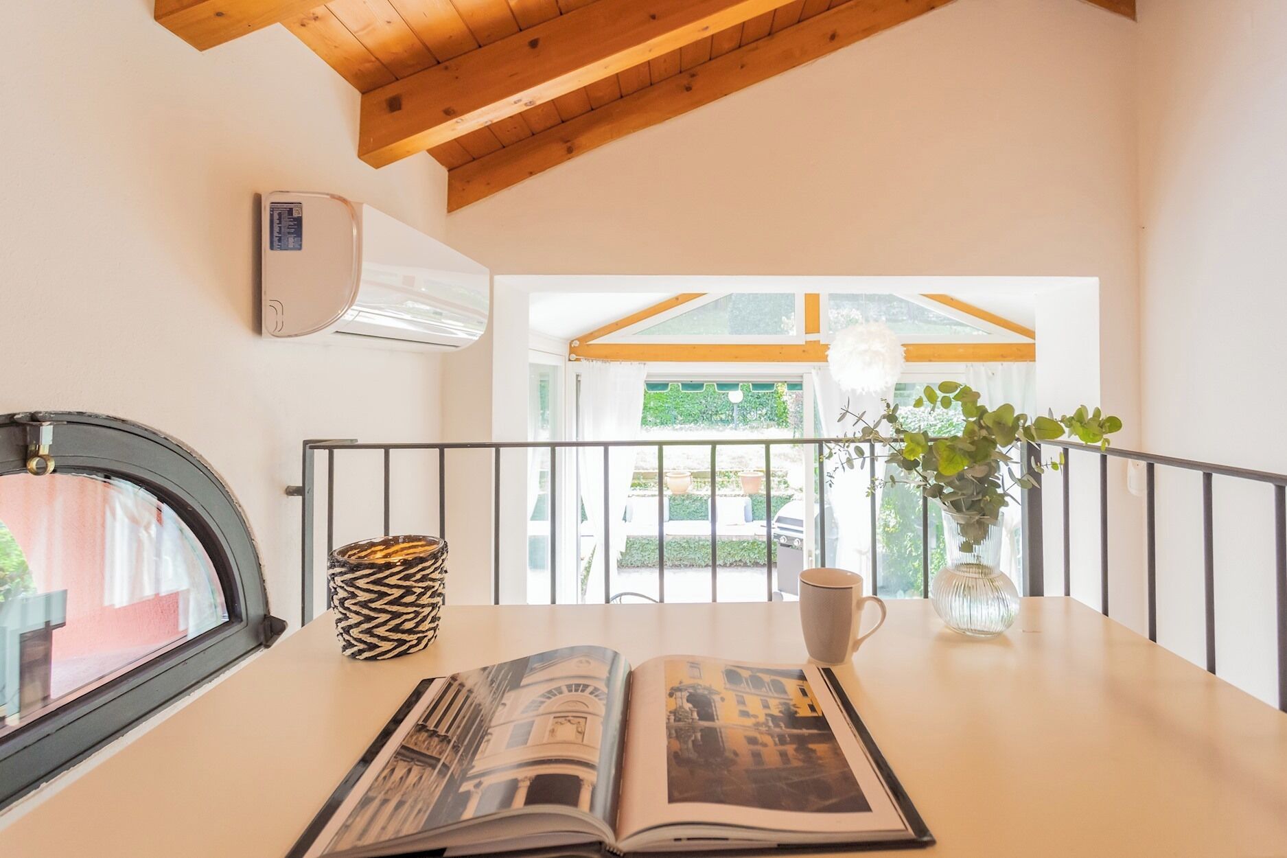 A room with a wooden ceiling, a window with a view of greenery, a table with a magazine, a coffee cup, and a decorative vase.