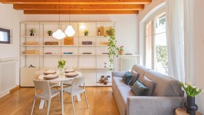 A modern living room with a dining area, wooden ceiling beams, a sofa, a small table with a plant, and a bookshelf with various items. - Vico Garden-203 by Rent all Como (Como)
