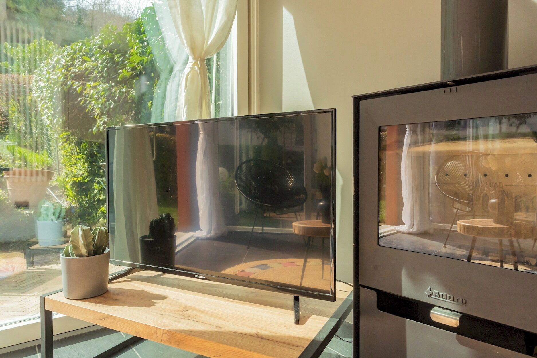 A modern living room with a large flat-screen TV on a wooden stand, a fireplace, and a view of a garden through the window.