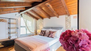 A bedroom with a wooden ceiling, a stone wall, a bed with a pink blanket, and a nightstand with a lamp.