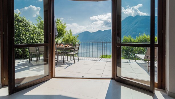 A room with a large sliding glass door leading to an outdoor terrace with a table and chairs, overlooking a mountainous landscape.