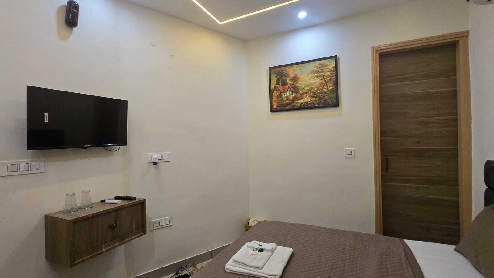 A hotel room with a bed, a TV mounted on the wall, a painting above the bed, a wooden door, and a small wooden cabinet with a mirror.