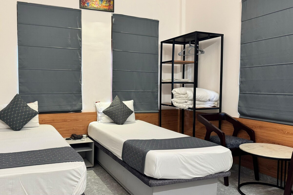 A hotel room with two single beds, a small table, a chair, and a shelf with folded towels.