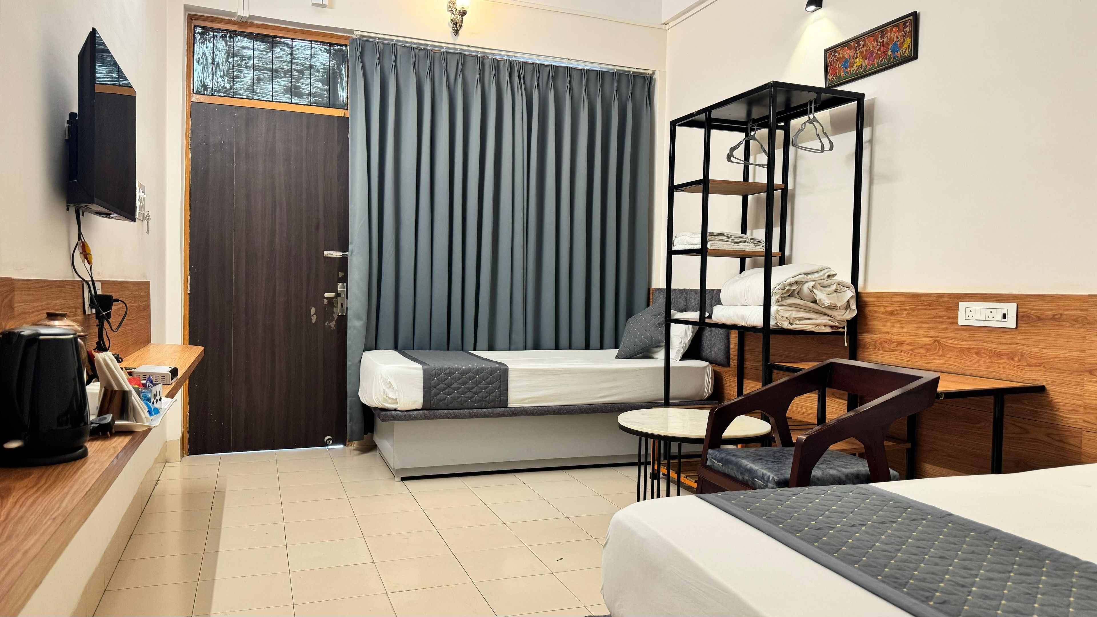 A compact hotel room with a bed, a small desk, a chair, a shelf with hanging clothes, and a television mounted on the wall.