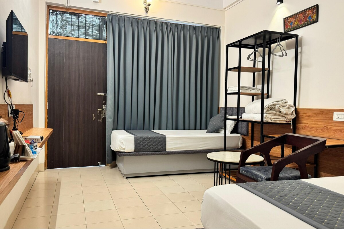 A compact hotel room with a bed, a small desk, a chair, a shelf with hanging clothes, and a television mounted on the wall.