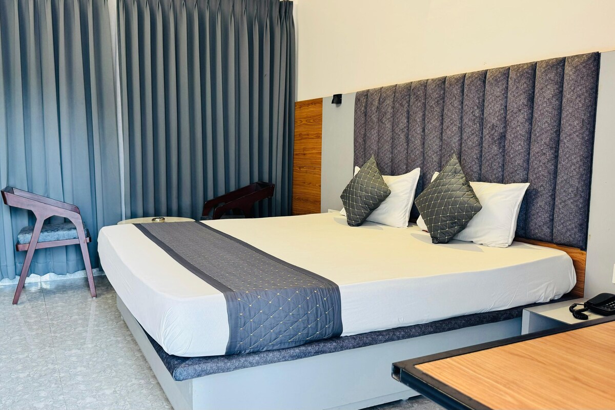 A hotel room with a large bed, a wooden headboard, grey curtains, a small wooden nightstand, and a telephone.