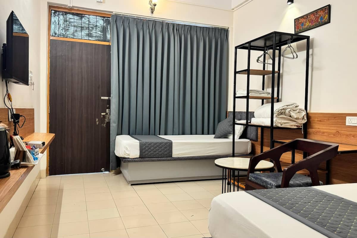 A compact hotel room with a bed, a small desk, a chair, a shelf with hanging items, and a television mounted on the wall.