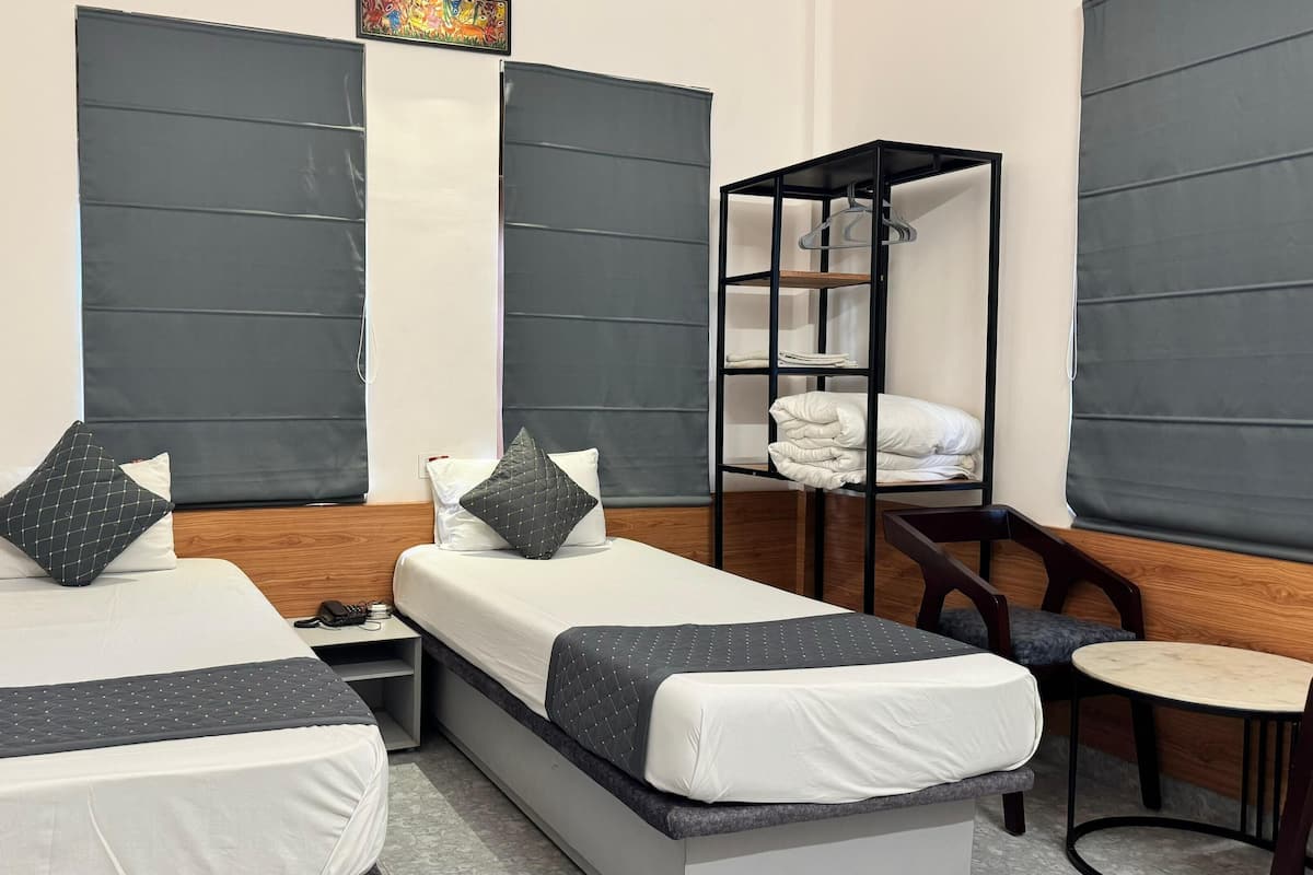 A hotel room with two single beds, a small table, a chair, and a shelf with folded blankets.