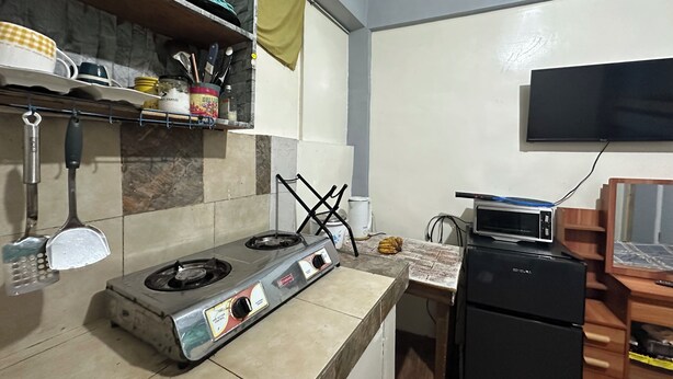 Eine Küche mit Gasherd, Mikrowelle und kleinem Kühlschrank. Ein Regal mit Küchenutensilien und ein an der Wand befestigter Fernseher.