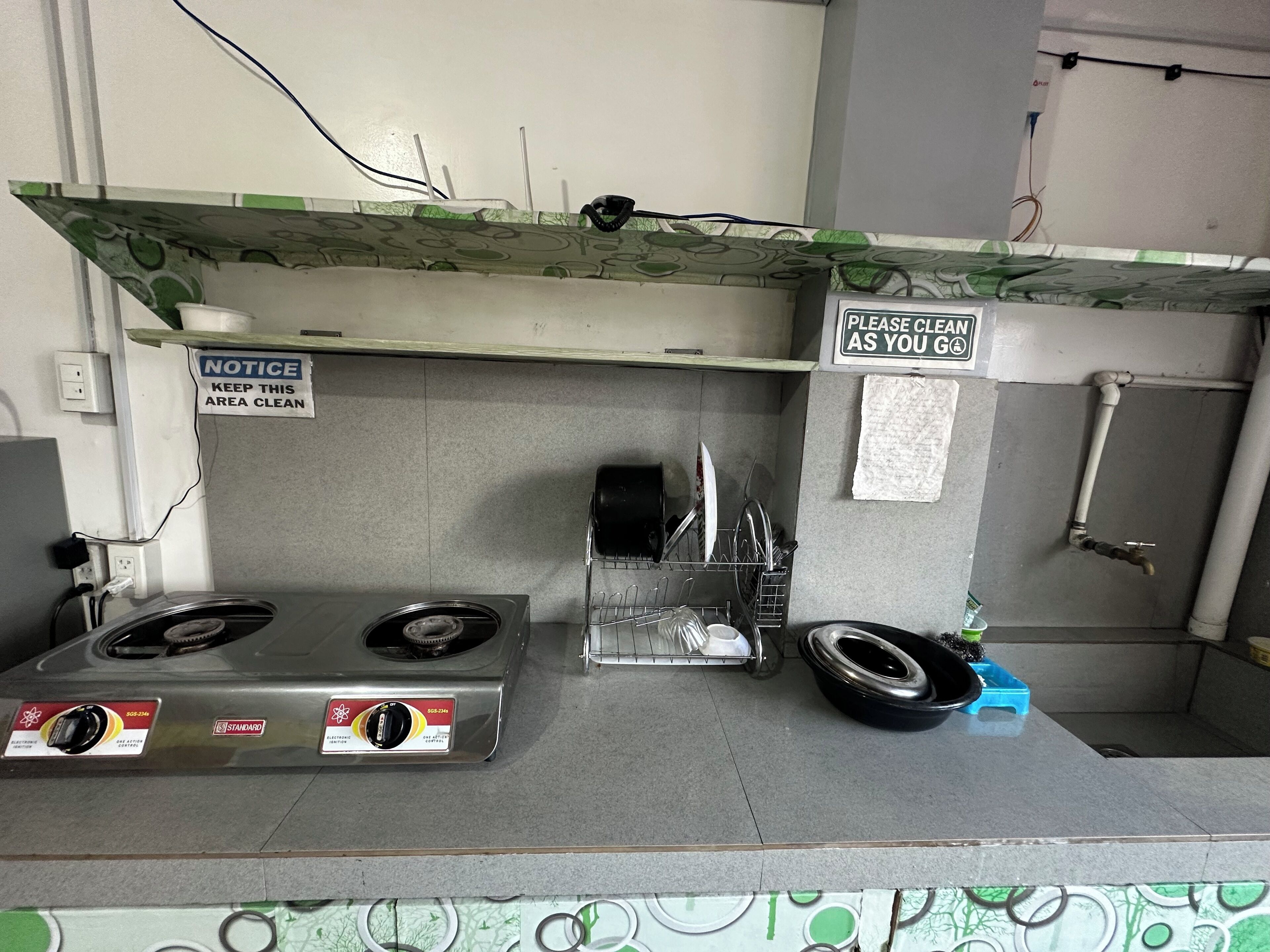 Une petite cuisine équipée de deux plaques de cuisson à gaz, d’un égouttoir et d’une pancarte rappelant de maintenir les lieux propres.