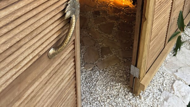 Une porte en bois avec une poignée en métal, qui donne sur une pièce au sol en pierre, avec une petite étagère où sont posées des bouteilles et une plante.