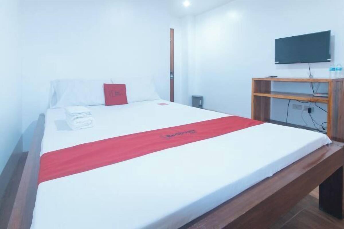 A hotel room with a bed, a television, a wooden bench, and a red bedspread.
