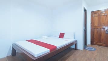 Una cama individual con un detalle rojo, una puerta de madera y una alfombra pequeña sobre piso de madera.