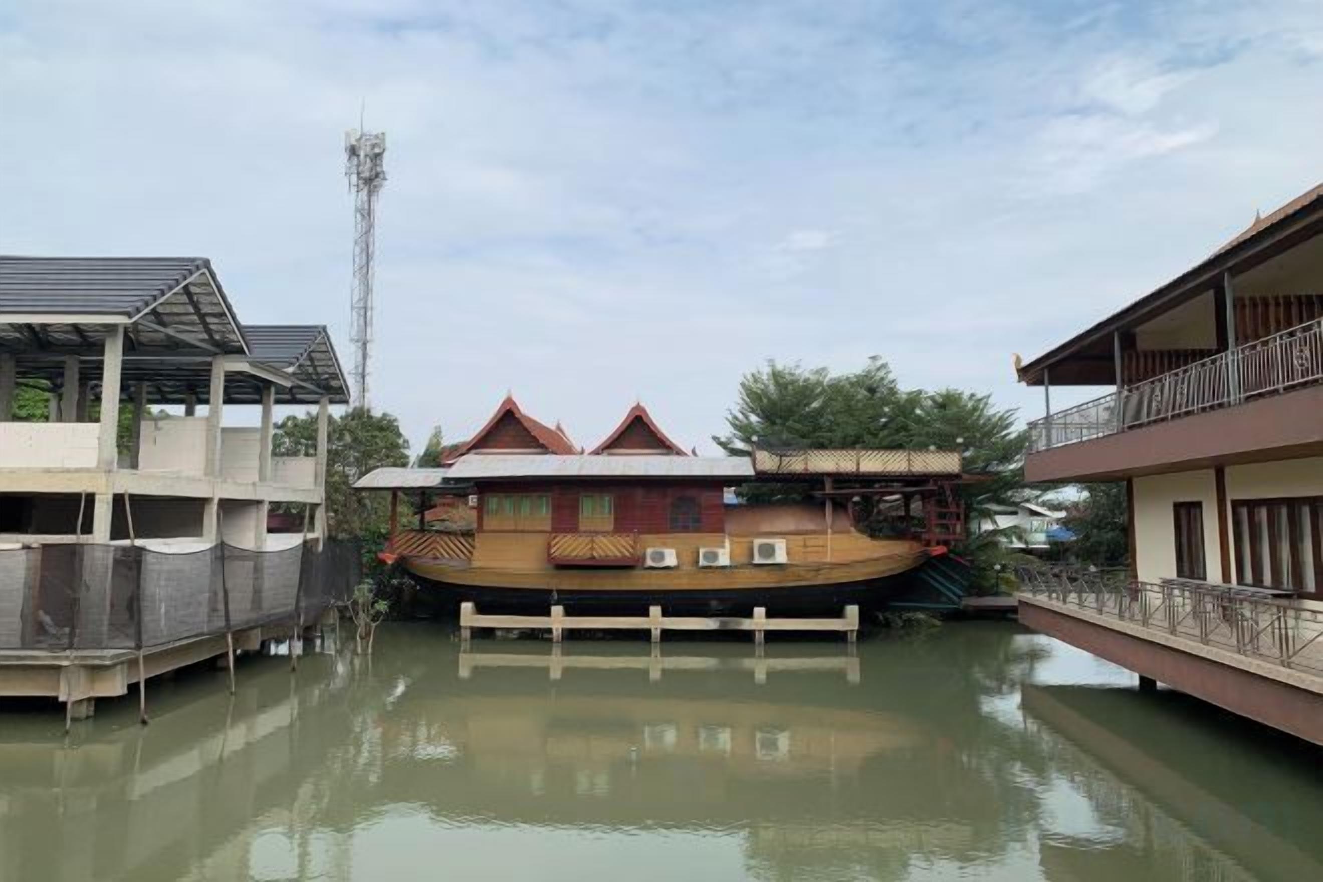 A traditional houseboat with a balcony, docked in a calm water body, surrounded by other structures.