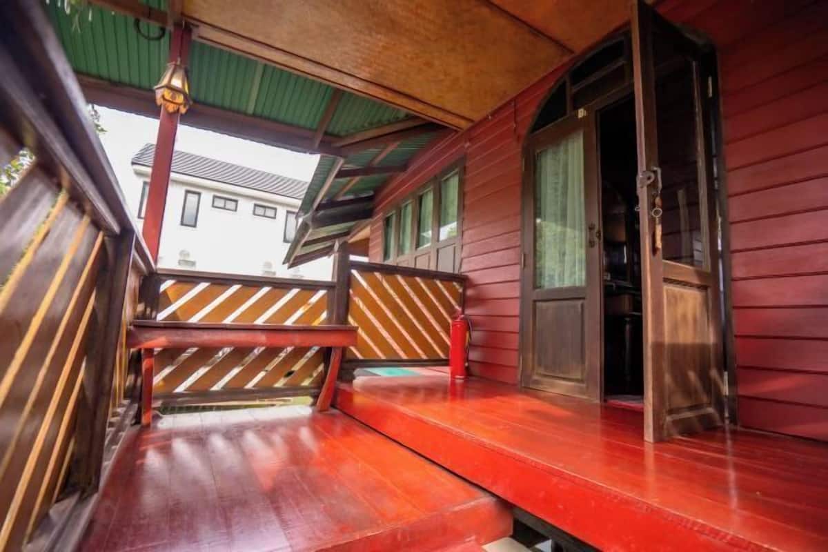Varanda de madeira com um banco, uma porta e vista para um edifício ao fundo.