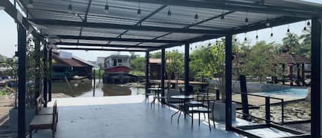 An outdoor covered patio area with tables and chairs, a view of a river, and a distant building.