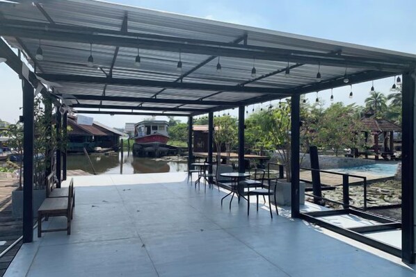Un área de patio techada al aire libre con mesas y sillas, vista a un río y un edificio distante.