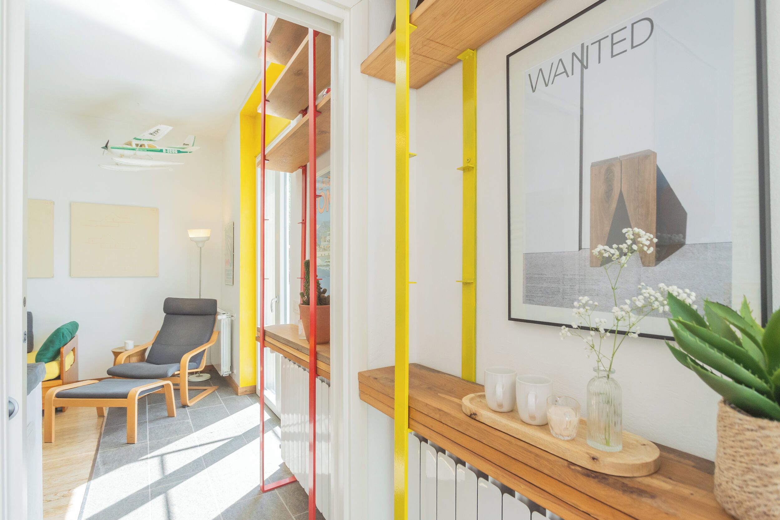 Een modern interieur met een houten bankje, een stoel, een plant en een 'WANTED' poster aan de muur.