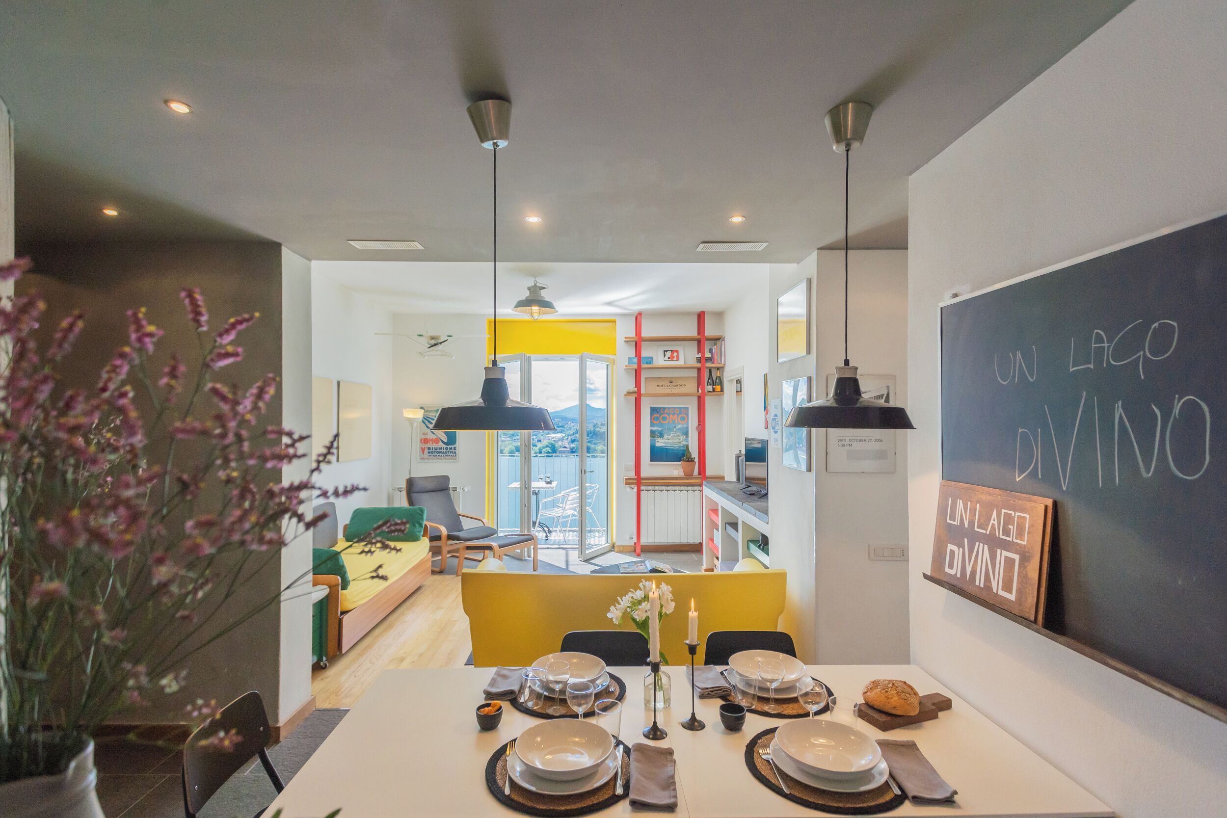 A modern dining area with a table set for four, a blackboard with text, and a view of the city through large windows.