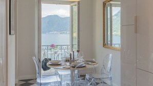 Comedor con mesa dispuesta para cuatro, sillas transparentes, una araña y vistas a un lago y montañas a través de un ventanal grande.